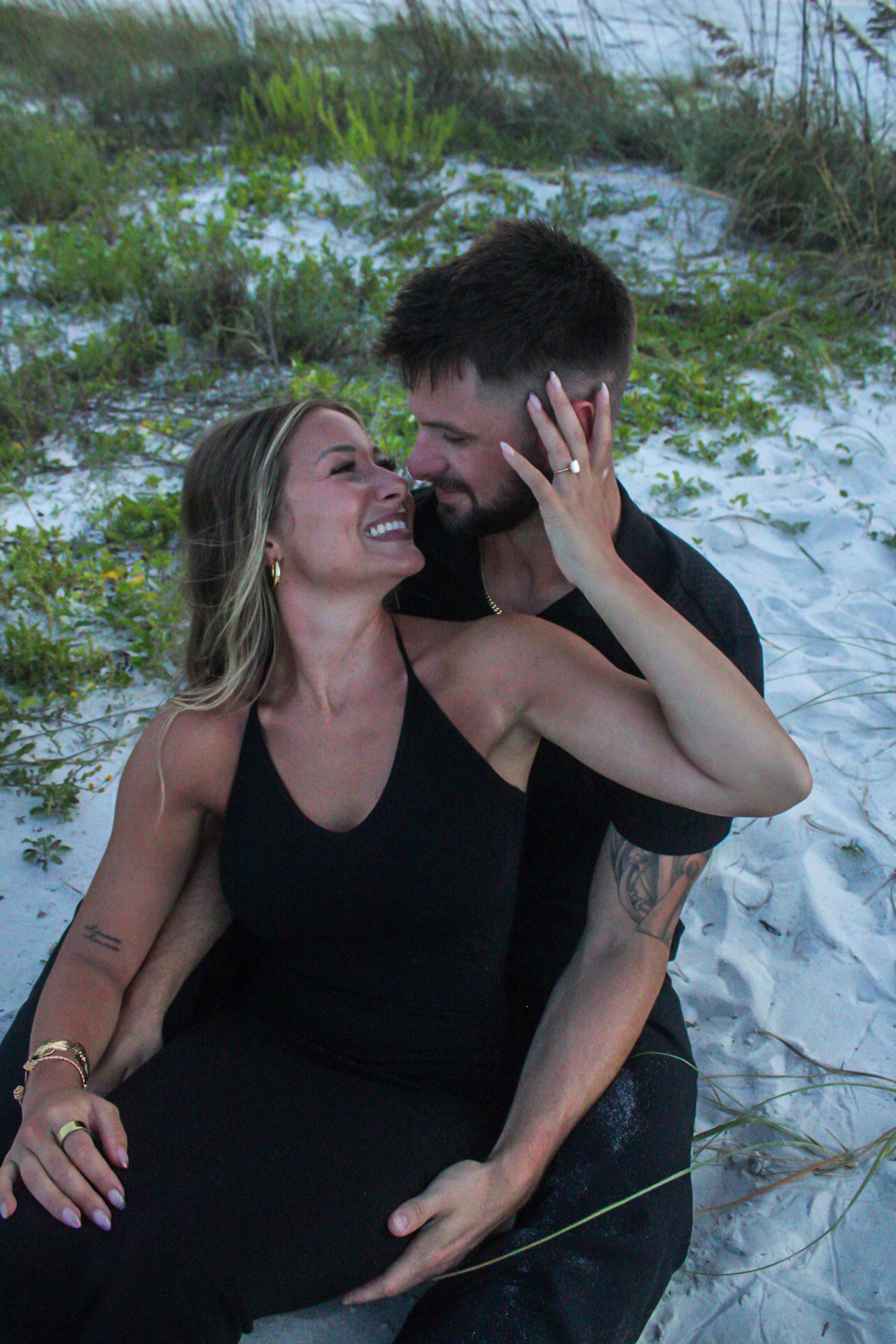 Couple dressed in black laughing together while seated in the sand near the dunes by Miranda Rae Photography