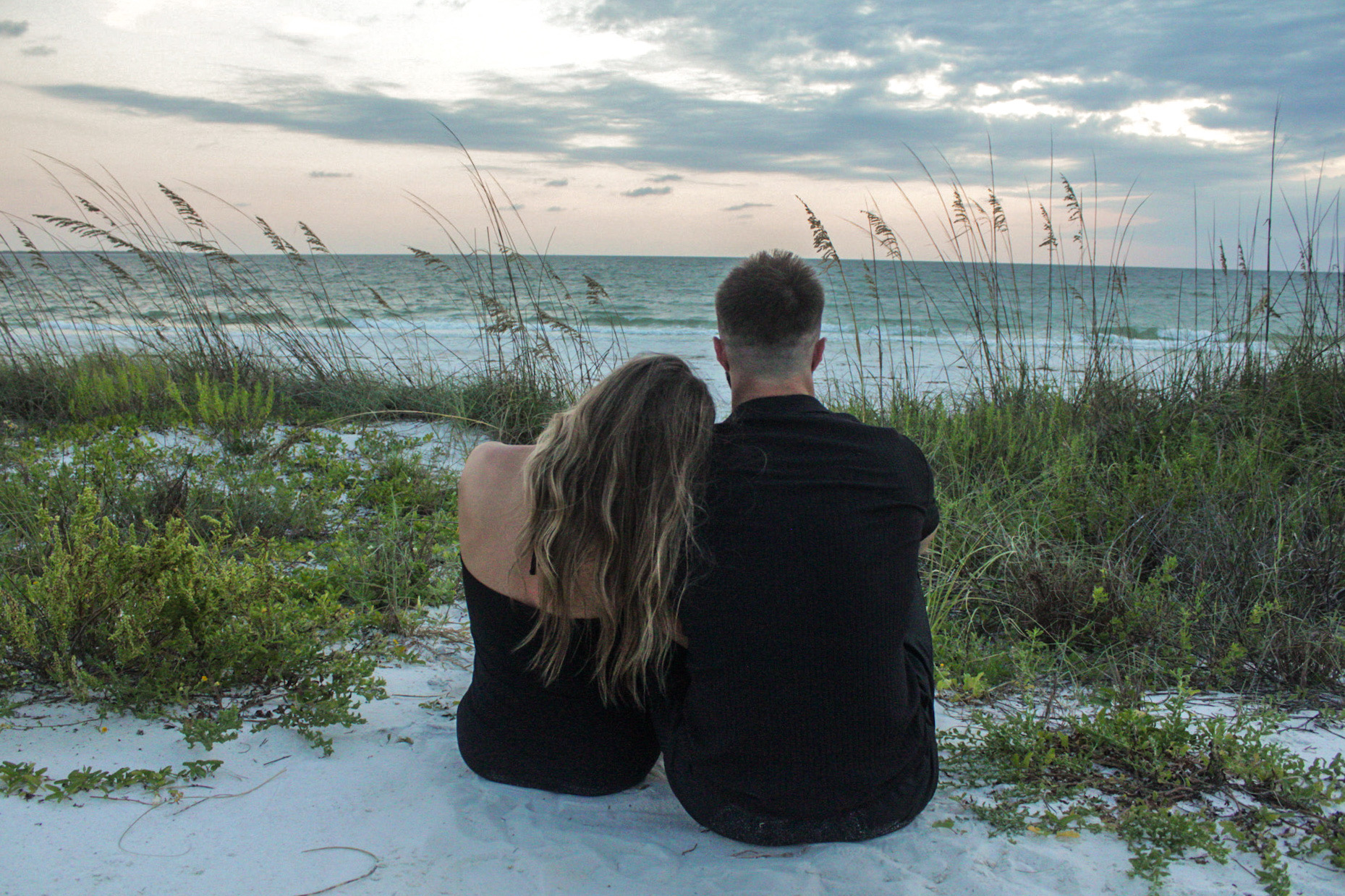 Couple seated together in the sand looking toward the ocean by Miranda Rae Photography